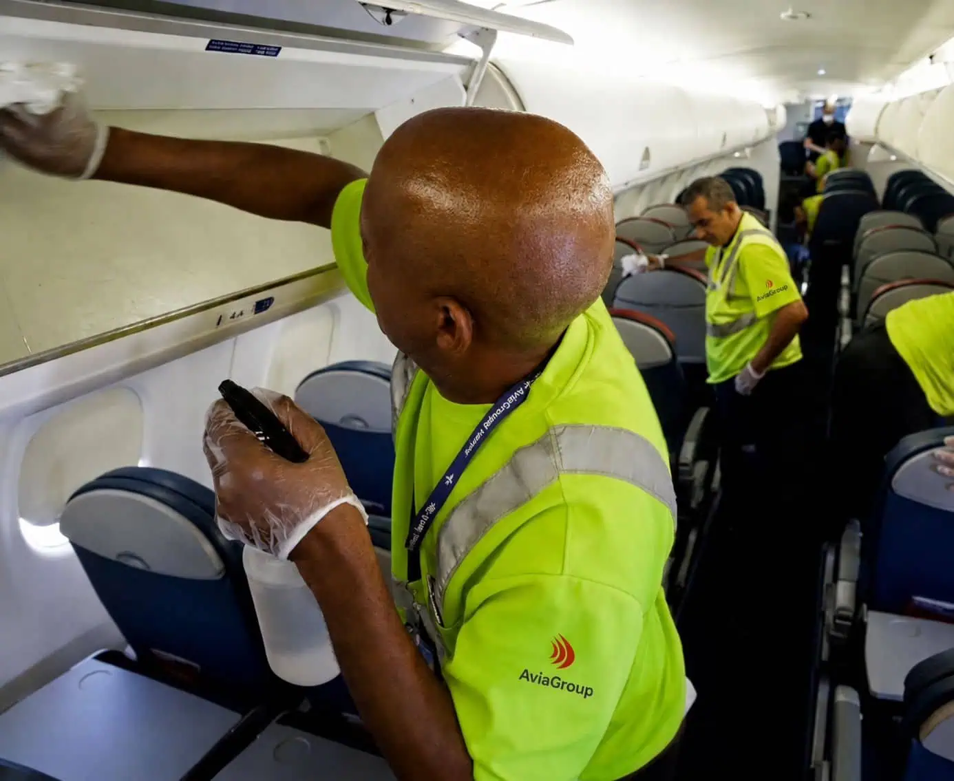 AviaGroup ground handling crew cleaning an aircraft cabin, wiping overhead bins and seats in hi-vis vests